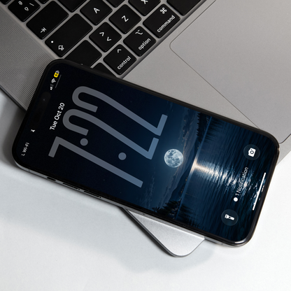 Phone resting on a laptop, displaying a full moon reflected in a dark blue lake between forested shores.