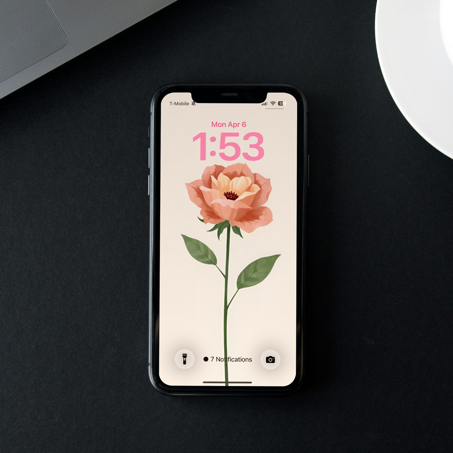 Smartphone with a floral screen on a dark surface next to a laptop and coffee cup.